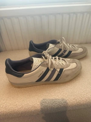 Adidas Gazelle vita och blå sneakers - Snygga Adidas Gazelle sneakers i vitt och ljusgrått med klassiska blå ränder på sidorna. Ovandelen är i mocka och läder, med vit plös och snörning. Låg modell som passar perfekt till jeans eller shorts. Ikonisk retrostil med diskret gulddetalj på sidan.