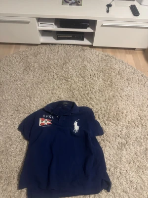 Marinblå piké från Ralph Lauren - Säljer en marinblå pikétröja från Ralph Lauren med klassisk krage, vita broderade detaljer och tryck på bröstet. Tillverkad i mjuk bomull och har korta ärmar. Perfekt för en stilren och sportig look.