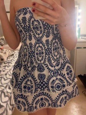 Vit och blå axelbandslös kortklänning - Säljer en söt axelbandslös kortklänning i vitt med blått broderat mönster. Klänningen har volangdetaljer och sitter löst på ett skönt sätt men ändå på plats. Beställde hem men gillade ej den på just mig, men tycker den är så fin. Aldrig använd. Orginalpris: 130kr
