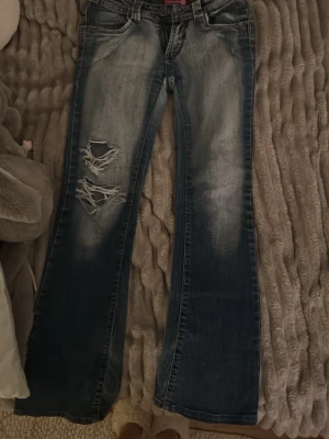 Blå bootcut jeans med slitningar - Säljer ett par blå bootcut jeans från Sabrina med slitningar och hål på vänster lår. Jeansen har ljusa tvättade partier, klassiska bakfickor med detaljer och en liten patch ovanför ena fickan. 