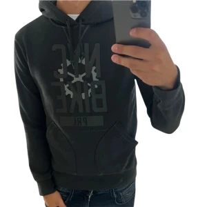 Svart hoodie från Ralph Lauren - Svart hoodie från Ralph Lauren med tryck 'NYC BIKE PRL' och coolt cykelhjulsmotiv på bröstet. Tröjan har huva med snörning och en stor magficka. Perfekt för dig som gillar streetstyle och vill ha en bekväm och snygg look.