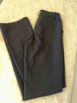 Svarta vida kostymbyxor - Säljer ett par svarta kostymbyxor med raka, vida ben och klassiska pressveck. Byxorna har hällor för skärp och diskreta bakfickor. Perfekta för en clean och stilren look. Materialet känns mjukt och följsamt.