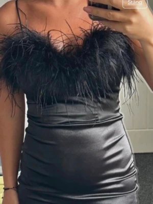 Svart satin klänning med fjädrar - Supersnygg svart klänning i satin med tight passform och fjäderdetaljer upptill. Klänningen är axelbandslös och har en kort längd som ger en trendig look. Perfekt för dig som vill sticka ut och känna dig extra glam.