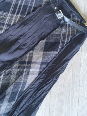 Svart långkjol med bälte och rutigt mönster - Snygg svart långkjol med rutiga paneler i grå och vita toner. Kjolen har ett tunt, glansigt material och ett svart bälte med silverfärgat spänne i midjan. Perfekt för dig som gillar en unik och cool stil.