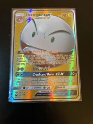 Electrode GX Pokémonkort holografiskt - Säljer ett Electrode GX Pokémonkort med holografisk yta. Kortet har guldfärgad ram och motiv av Electrode med starka attacker som Extra Energy Bomb, Electro Ball och Crush and Burn GX. Perfekt för samlare eller Pokémon TCG-spelare.