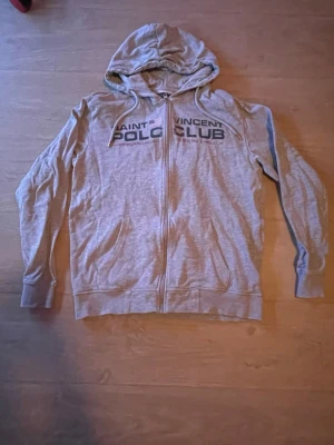 Grå Saint Vincent Polo Club hoodie - Snygg grå hoodie med dragkedja från Saint Vincent Polo Club. Tryck på bröstet och två fickor framtill. Tillverkad i mjukt material, perfekt för en avslappnad stil. Klassisk huva med dragsko och ribbade muddar vid ärmar och nederkant.