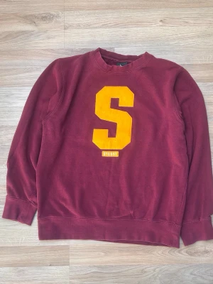 Vinröd sweatshirt från Stussy - Cool vinröd sweatshirt från Stussy med stor gul S på bröstet. Klassisk crewneck-modell med ribbade muddar vid ärmar och nederkant. Perfekt för dig som gillar streetwear och vill sticka ut lite extra.