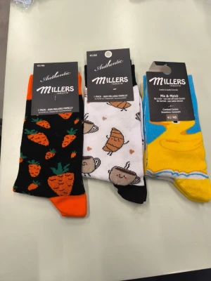 Färgglada strumpor från Millers - Tre par strumpor från Millers med olika motiv: ett par med morötter på svart botten, ett par vita med croissanter och kaffekoppar, samt ett par med frukostmotiv i gult och blått. Strumporna är tillverkade i bomull och har roliga, färgstarka mönster.