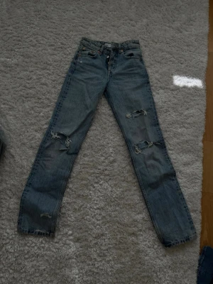Blå slitna raka midrise jeans - Säljer ett par blå jeans med raka ben och flera slitningar på låren och knäna. Jeansen har klassisk femficksdesign och är gjorda i denim. Perfekta för en avslappnad och trendig look.