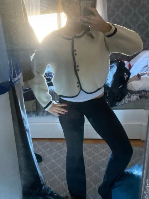 Vit kofta med svarta detaljer - Säljer en söt vit kofta med svarta kanter och dekorativa knappar framtill. Koftan har rundad krage, långa ärmar och en lite boxig, croppad passform. Perfekt att styla till både jeans och kjol för en trendig look.