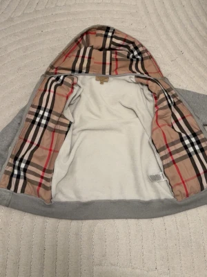 Grå hoodie från Burberry med mönstrad huva - Säljer en grå hoodie från Burberry med den klassiska beigea, röda och svarta rutiga insidan på huvan och fodret. Hoodien är mjuk och har en riktigt lyxig känsla med sitt ikoniska Burberry-mönster. Perfekt för dig som vill ha något stilrent men ändå sticker ut.