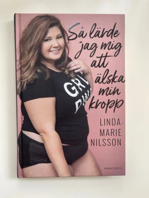 Så lärde jag mig att älska min kropp - En inspirerande bok av Linda-Marie Nilsson om kroppspositivism och självacceptans. Omslaget är rosa med svart text och ett foto på författaren. Inbunden bok som tar upp resan till att älska sig själv och bryta normer kring kroppsideal. Helt ny och oläst!