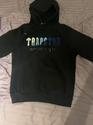 Trapstar hoodie - Säljer en svart hoodie från Trapstar med stor huva och dragsko. På bröstet står det 'TRAPSTAR' i gradient från vitt till blått och texten 'IT'S A SECRET' under. Klassisk känguruficka framtill och ribbade muddar. Perfekt för en streetwear-look.