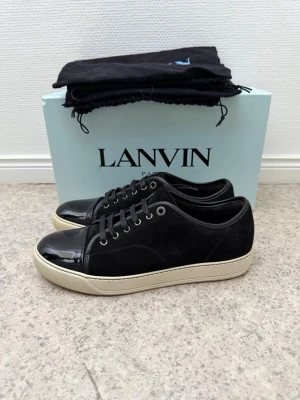 Svarta Lanvin sneakers med lackdetalj - Snygga svarta sneakers från Lanvin med klassisk låg profil. Skorna har en unik kombination av mocka och glansig lack på tån, samt vita sulor som ger en stilren kontrast. Perfekta för dig som vill ha en exklusiv och trendig look. Uk8. Dustbag och skor medföljd endast