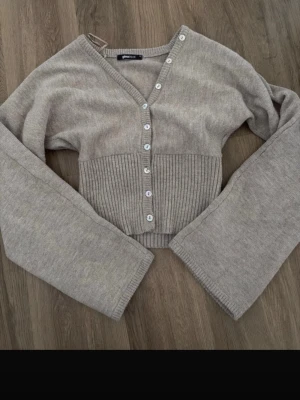 Beige kofta från Gina Tricot - Kort beige kofta från Gina Tricot med v-ringning och pärlemorknappar framtill. Koftan har bred ribbstickad midja och långa, vida ärmar. Perfekt att styla med höga jeans eller kjol för en trendig look.