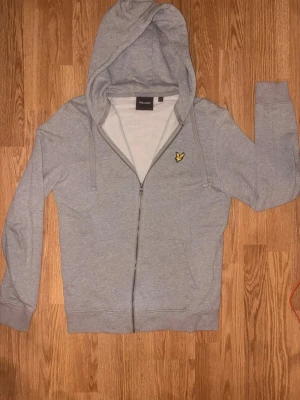 Grå zip hoodie från Lyle & Scott - Snygg grå hoodie med dragkedja från Lyle & Scott. Klassisk design med huva och dragsnören, samt den ikoniska gula fågel-loggan på bröstet. Tillverkad i mjukt bomullsmaterial och har ribbade muddar vid ärmar och nederkant. Perfekt för en avslappnad stil.