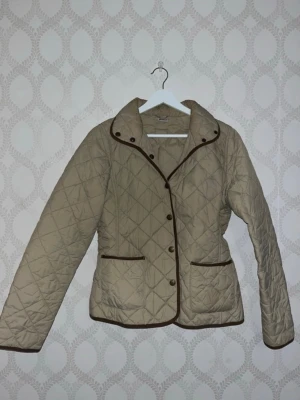 Beige quiltad jacka med bruna detaljer - Stilren beige quiltad jacka med bruna kantband och knappar framtill. Jackan har två fickor och en klassisk krage. 