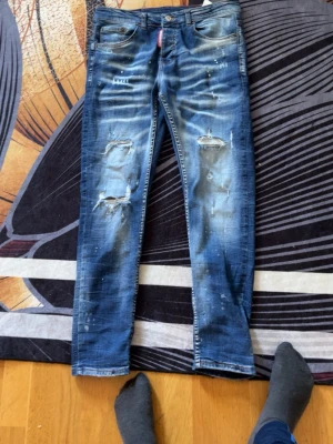 Blå slitna jeans från Dsquared2 är äkta  - Säljer ett par blå jeans från Dsquared2 med slitningar och hål på benen samt coola färgstänk. Jeansen har en smal passform och klassisk femficksdesign. Perfekta för dig som gillar en edgy och trendig stil.