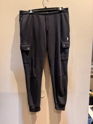 Svarta cargo-mjukisbyxor Polo Ralph Lauren - Svarta mjukisbyxor från Polo Ralph Lauren med cargo-fickor på sidorna och broderad logga på vänster ben. Byxorna har muddar vid bensluten och knappdetaljer på fickorna. Tillverkade i mjukt sweatshirtmaterial för maximal komfort.
