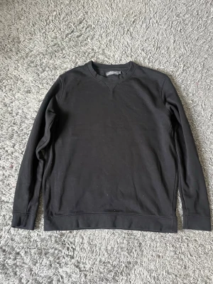 Svart sweatshirt från Dressmann - En klassisk svart sweatshirt från Dressmann i storlek L. Tröjan har rund halsringning och långa ärmar, perfekt för en enkel och stilren look. Tillverkad i mjuk bomull för skön komfort hela dagen.