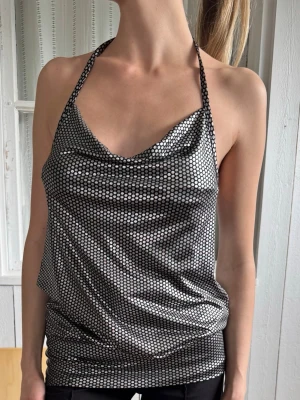 Silvrig halterneck topp med nätmönster - Snygg silvrig halterneck topp med nätmönster och draperad front. Toppen är ärmlös och har öppen rygg, perfekt för dig som vill sticka ut. Materialet känns glansigt och ger en cool look.