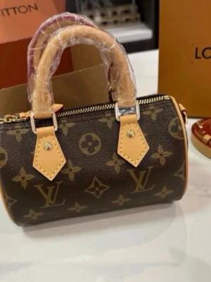 Louis Vuitton väska - Snygg brun handväska från Louis Vuitton med klassiskt monogrammönster och beige detaljer. Väskan har två korta handtag och guldfärgade metalldetaljer. Perfekt accessoar för dig som gillar lyxig och tidlös design.
