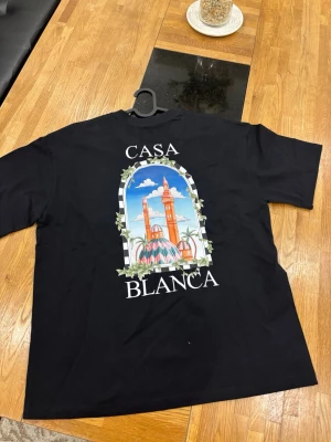 Svart Casablanca t-shirt med tryck - Svart t-shirt från Casablanca med stort färgglatt tryck av byggnader och palmer på ryggen samt mindre tryck på bröstet. Klassisk passform och rund hals. Perfekt för dig som vill ha en stilren men ändå unik look.