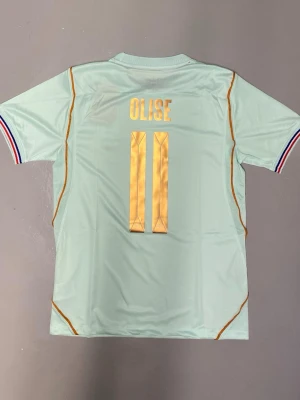 Olise#11 VM 2026 Frankrike fotbollströja Olise - Snygg ljusblå fotbollströja med Olise och nummer 11 i gult på ryggen. Tröjan har korta ärmar med ränder i blått, vitt och rött samt tunna orange detaljer längs sidorna. Frankrikes landslagsemblem och Nike-logga framtill.