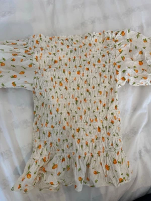 Vit blommig topp H&M - Supersöt vit topp från H&M med smockad kropp och volangkant. Toppen har korta puffärmar och ett gulligt blommigt mönster i orange och grönt. Perfekt för vår och sommar, lätt och luftig i bomull. Aldrig använd. Storlek M passar ner till XS/XSS och upp till L då den är stretchig. 