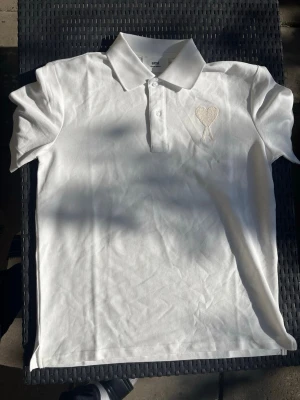 Vit piké med hjärtdetalj - Snygg vit pikétröja med krage och knappar framtill. På bröstet finns en broderad hjärtdetalj i samma färg som tröjan. Perfekt för en clean och stilren look.