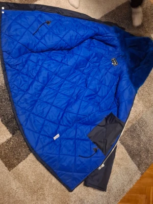 Mörkblå parkas med blått foder - Snygg mörkblå parkas med quiltat, starkt blått innerfoder och luva med fuskpäls. Jackan har flera stora fickor framtill, dragkedja och knappar samt en ficka på ärmen. Perfekt för kalla dagar tack vare det tjocka fodret och den höga kragen.