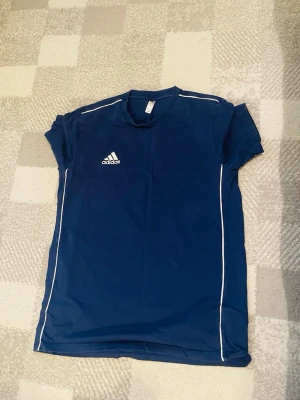 Blå sporttröja från Adidas - Mörkblå sporttröja från Adidas med vita detaljer längs axlar och sidor. Tröjan har rund halsringning och korta ärmar. Adidas-loggan är tryckt på bröstet. Perfekt för träning eller fotboll.
