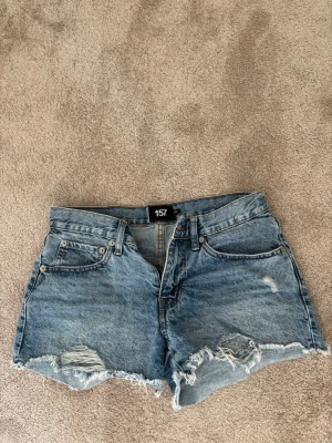 Blå denimshorts från lager 157 - Säljer ett par blå denimshorts från 157 med låg midja och råa, slitna kanter. Shortsen har klassiska fickor, bälteshällor och dragkedja med knapp. Perfekta för sommaren och ger en avslappnad vibe.