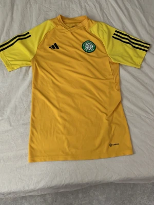 Gul Adidas Celtic träningströja - Säljer en gul träningströja från Adidas med Celtic FC-logga på bröstet. Tröjan har ljusgula ärmar med svarta Adidas-ränder och är tillverkad i ett lätt, snabbtorkande material. Perfekt för fotbollsträning eller gymmet.