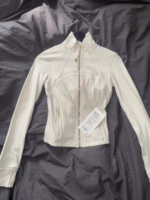 Lululemon define jacket cropped Nulu. Strl 4 (XS) - Snygg vit jacka från lululemon med guldfärgade dragkedjor och hög krage. Figurnära passform och långa ärmar. Tillverkad i ett stretchigt material som ger en sportig och clean look. Strl 4 som jag skulle uppskatta till XS. Aldrig använt!