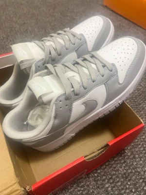 Nike Dunk Low gråvita sneakers - Nike Dunk Low sneakers i vitt och ljusgrått med klassisk Swoosh på sidan. Ovandelen är i slätt läder med perforerad tåbox för extra ventilation. Lågt skaft och matchande grå skosnören ger en clean och stilren vibe.