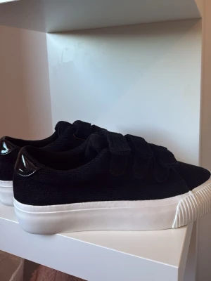 Svarta sneakers med kardborreband från EVEN&ODD - Säljer ett par oanvända svarta sneakers med breda kardborreband och vit tjock sula från EVEN&ODD. Skorna har en ovandel i manchestertyg och en detalj i blankt material vid hälen. Perfekta för dig som gillar en clean och stilren look.