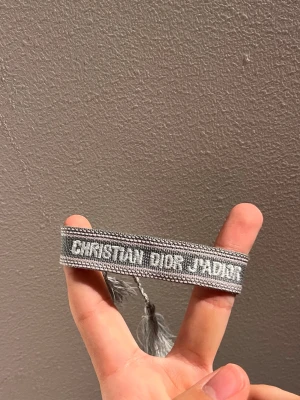 Grått armband Christian Dior - Snyggt grått tygarmband från Christian Dior med texten 'CHRISTIAN DIOR J'ADIOR' broderad i vitt. Armbandet har dekorativa tofsar i ändarna och är justerbart för att passa olika handleder. Perfekt accessoar för att lyfta din outfit.