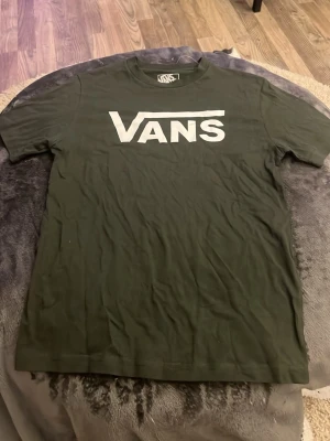 Svart Vans t-shirt med logga - Säljer en mörk grön t-shirt från Vans med stor vit logga tryckt över bröstet. Klassisk rund hals och korta ärmar. Den är skön att bära. Perfekt för dig som gillar streetwear och enkel stil.