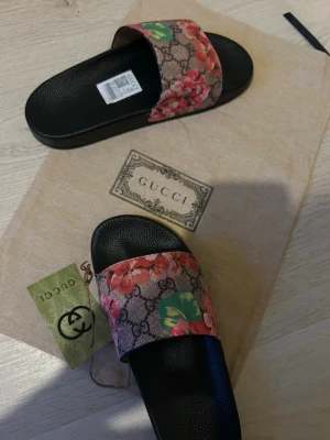 Sandaler - Snygga sandaler svart sula och bred rem i canvas med klassiskt GG-mönster och färgglada blommor i rosa, rött och grönt. Det är storlek 38 men väldigt små i storlek så passar 36-37. Medföljer dustbag