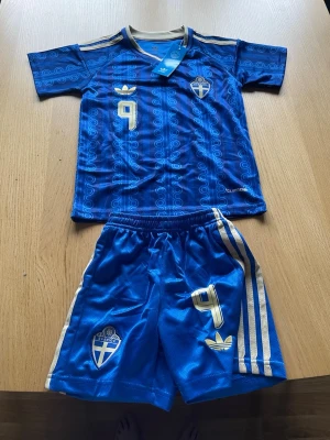 Blått Sverige fotbollskit Adidas - Säljer ett blått fotbollskit från Adidas med Sveriges landslagstryck. Tröjan har nummer 9 och ISAK på ryggen, samt guldiga detaljer och emblem. Matchande shorts med nummer 9 och Sveriges märke. Perfekt för fotbollsträning eller match. Passar till ålder runt 8 år