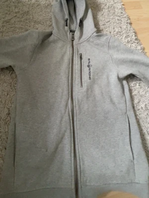 Grå hoodie från Sail Racing - Snygg grå hoodie från Sail Racing med dragkedja framtill och en extra dragkedjeförsedd bröstficka. Broderad logga på bröstet. Mjuk bomullsblandning och huva med dragsko. Perfekt för chill eller streetwear.