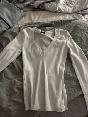 Vit ribbad långärmad topp - Snygg vit ribbad topp med lång ärm och djup v-ringning. Perfekt att styla med jeans eller kjol för en clean look. Materialet är stretchigt och sitter skönt mot kroppen.