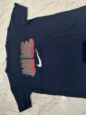 Mörkblå Nike t-shirt med tryck - Snygg mörkblå t-shirt med coolt grafiskt tryck i rött, grått och vitt på bröstet samt Nike-logga. Klassisk passform och rund halsringning. Perfekt för en avslappnad streetstyle-look.