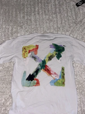 Off-white T-shirt - Vit off-white t-shirt med korta ärmar och färgstarkt konstnärligt tryck på ryggen i grönt, gult, rött, blått och svart. Framsidan har en liten tryckt detalj med text och färg. Perfekt för dig som gillar streetwear och unika plagg. Aldrig använt!! Känns som en storlek M!!