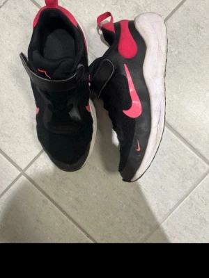 Svarta sneakers med rosa detaljer - Snygga svarta sneakers med rosa detaljer och vit sula. Skorna har en sportig design med kardborreband och snörning. Ovandelen är i mesh och syntetmaterial för bra andningsförmåga. Perfekta för dig som gillar en färgstark och sportig look.