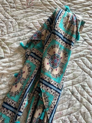 Mönstrad sjal med tofsar från DOCA - Säljer en sjal från DOCA med orientaliskt mönster i turkos, beige och marinblått. Sjalen har små turkosa tofsar längs kanterna och är tillverkad i ett tunt, mjukt tyg som känns skönt mot huden. Perfekt accessoar för att lyfta din outfit.