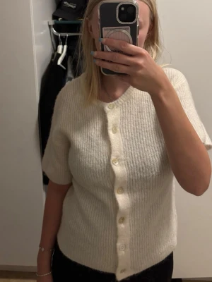Offwhite stickad kofta Gina Tricot - Mysig offwhite stickad kofta från Gina Tricot med korta ärmar och knappar framtill. Tillverkad i en mjuk ullblandning med 14% certifierad ull. Perfekt att slänga över en topp för en enkel och stilren look.