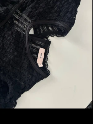 Svart blus med snörning från Nelly - Säljer en svart blus från Nelly med transparent rutigt mönster och fina spetsdetaljer. Toppen har snörning vid halsen och volang vid ärmsluten. Perfekt för dig som gillar trendiga och unika toppar.