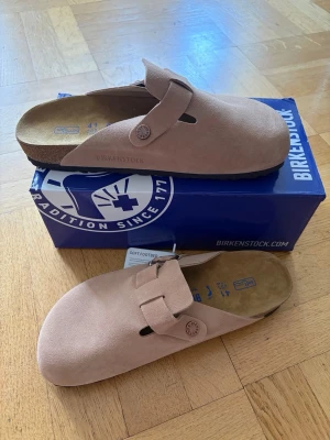 Birkenstock boston - Säljer ett par snygga  tofflor från Birkenstock i mjuk mocka. Modellen har en bred rem med spänne och klassisk korksula för extra komfort. Perfekta för dig som vill ha både stil och bekvämlighet hemma eller ute.